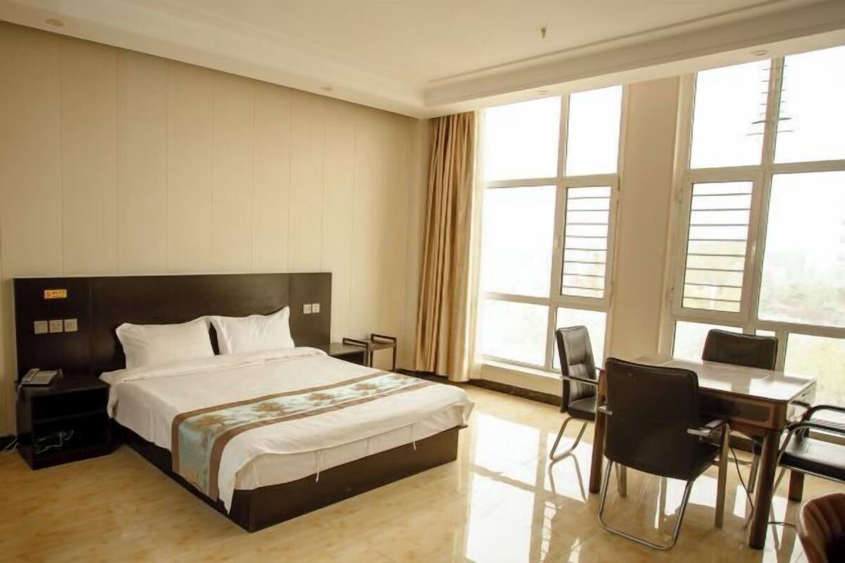 Habitación de hotel con una cama grande, un escritorio, sillas y un amplio ventanal con cortinas.