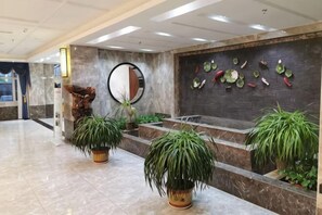 Vestíbulo con suelo de mármol, plantas en macetas, mostrador de recepción, espejo adosado a la pared y una pared decorativa con motivos de frutas.