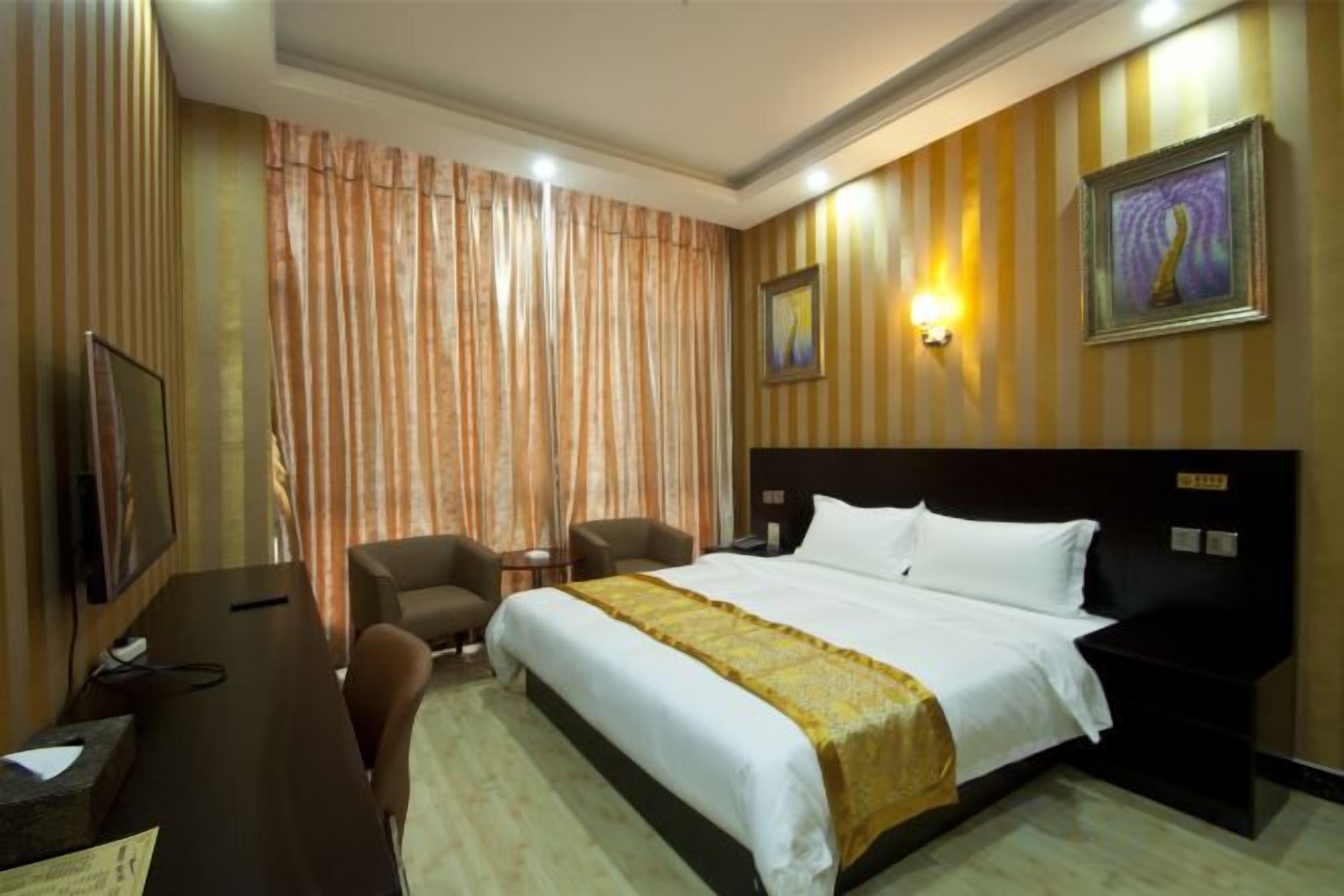 A hotel room with a bed, a desk with a chair, a television, and a wall-mounted lamp.