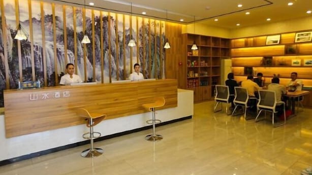 Een hotel receptie met een houten balie, twee barkrukken, een zithoek met tafels en stoelen, en een muurschildering van een waterval.