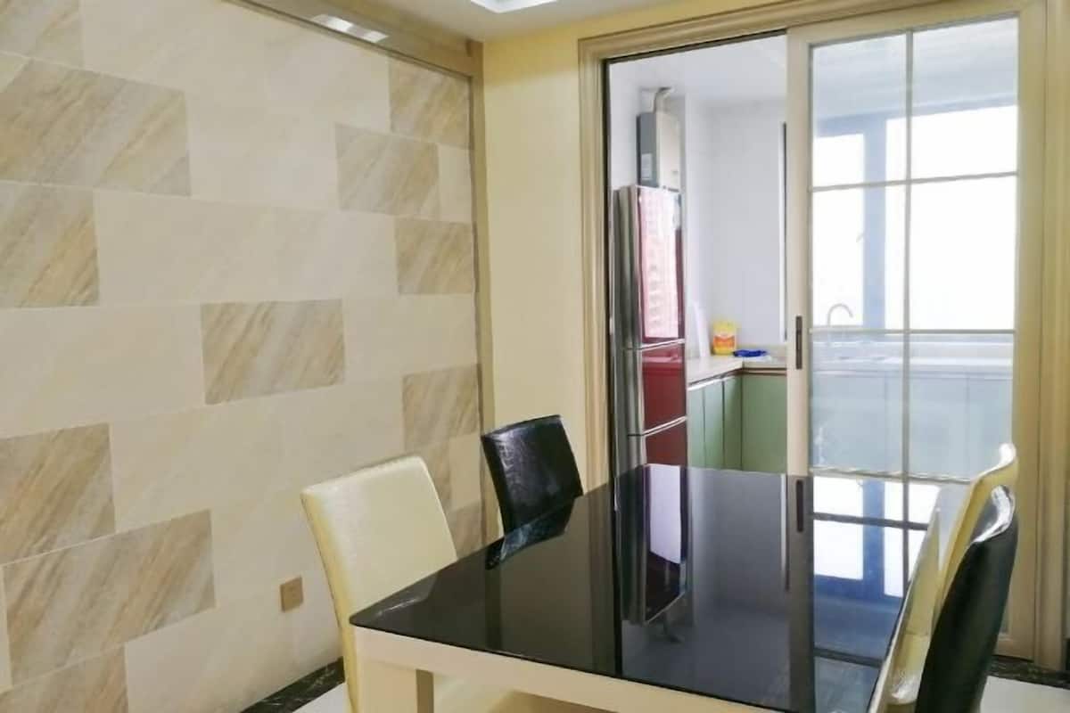 Een eetgedeelte met een zwarte tafel en witte stoelen, een betegelde muur, en uitzicht op een keuken en balkon door een glazen deur.