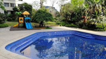 Una piscina con tobogán, rodeada de vegetación y un parque infantil.