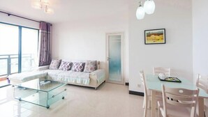 A modern living room with a glass coffee table, a sofa with patterned cushions, a dining table with chairs, and a framed picture on the wall.