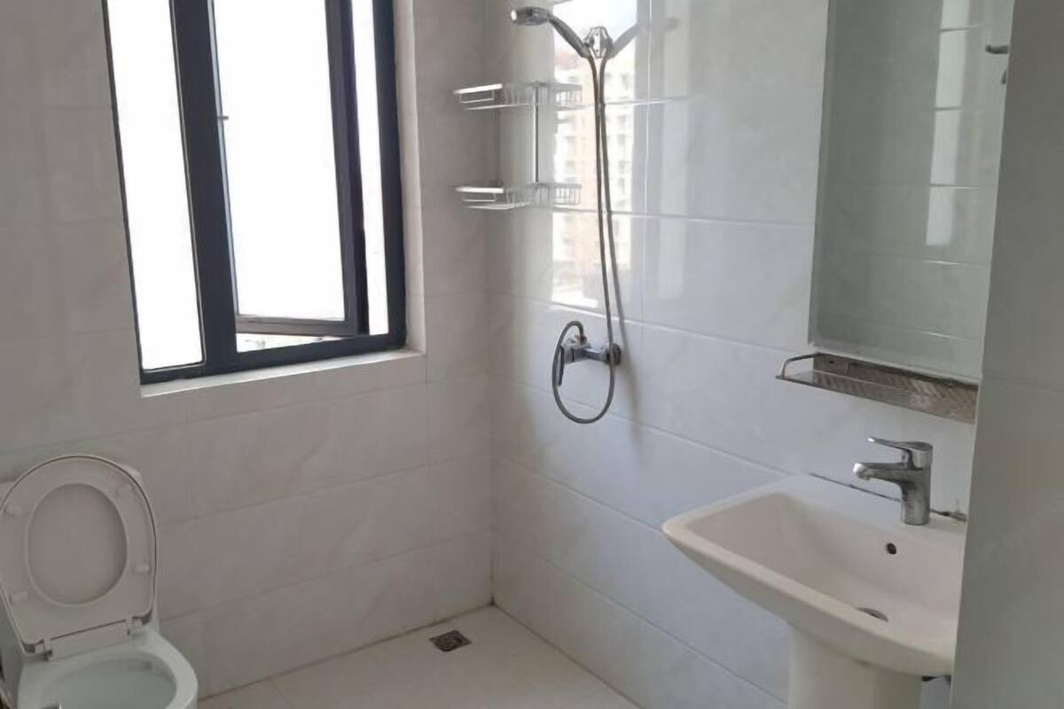 Een kleine, schone badkamer met witte tegels, een toilet, een wastafel, een douche met een handdouche en een raam voor natuurlijk licht.