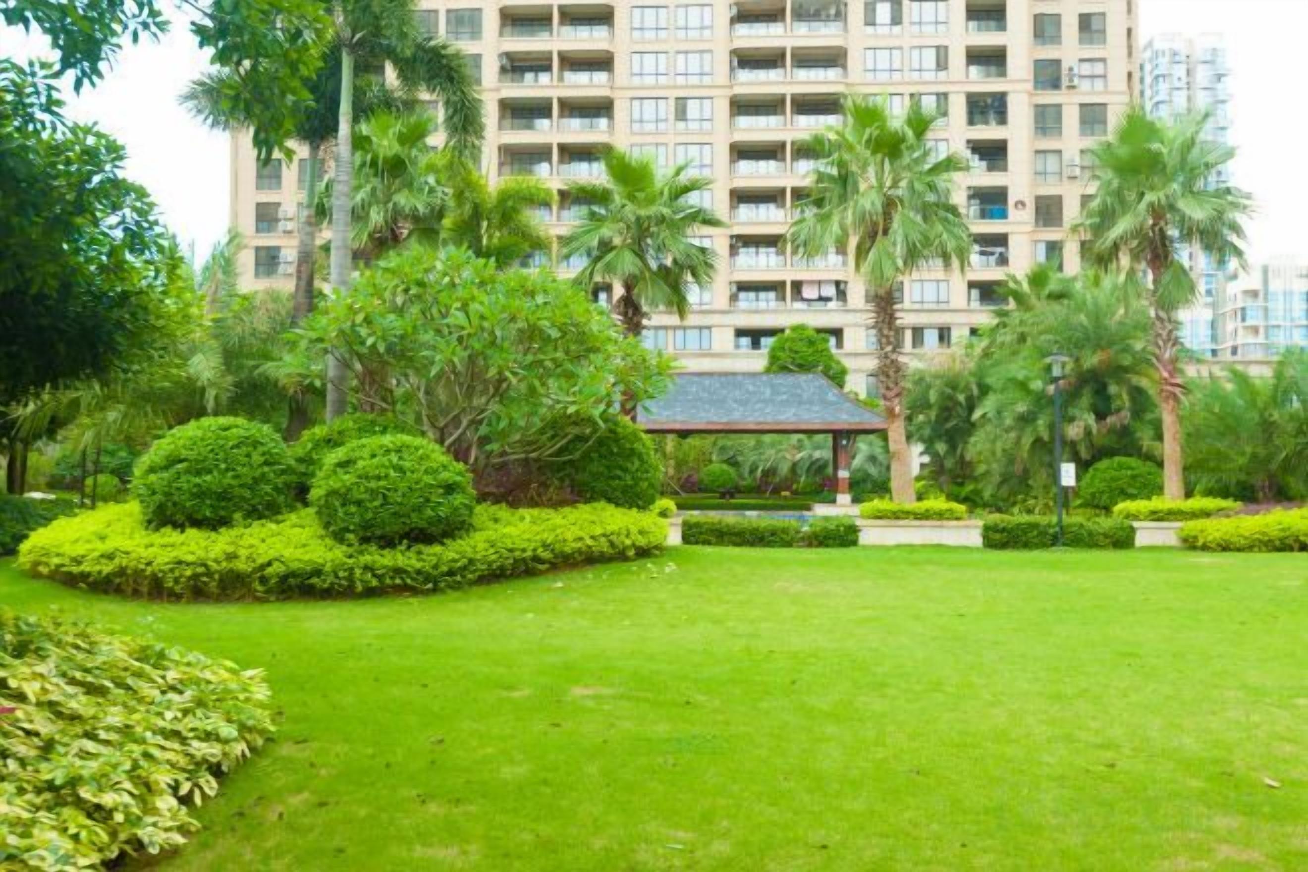 Un jardín bien cuidado con un pabellón, palmeras y un edificio de varios pisos al fondo.