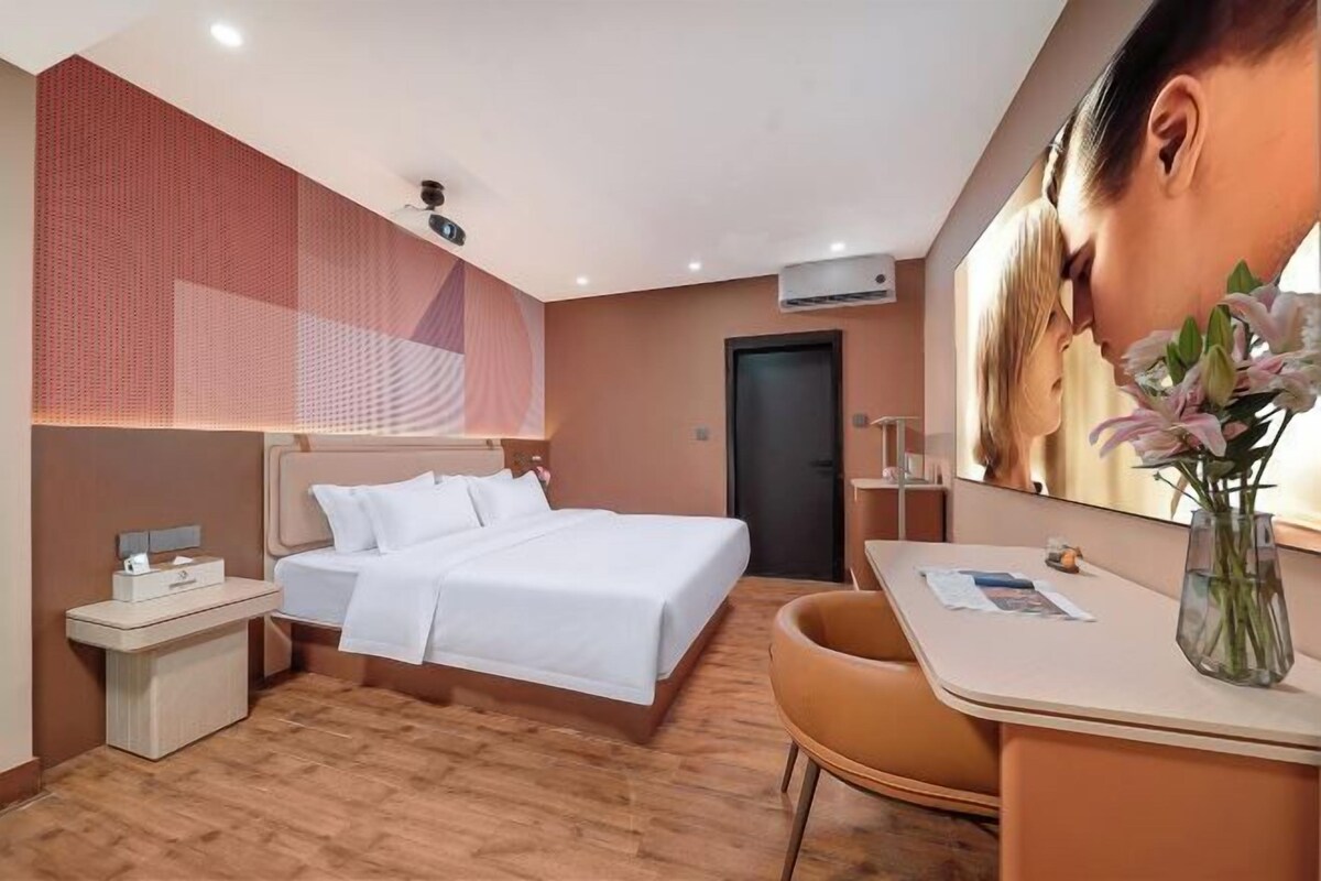 A hotel room with a bed, bedside table, chair, and a desk with a vase of flowers.