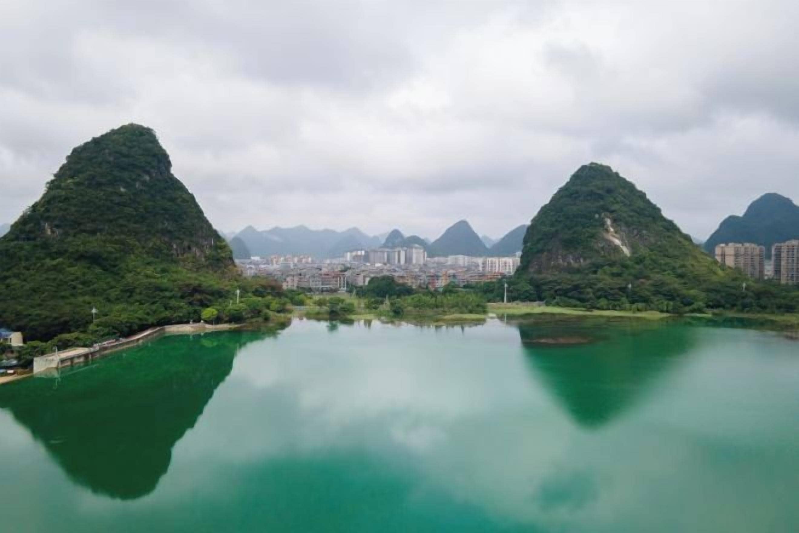Una superficie acuática tranquila que refleja dos colinas verdes, con un paisaje urbano al fondo.