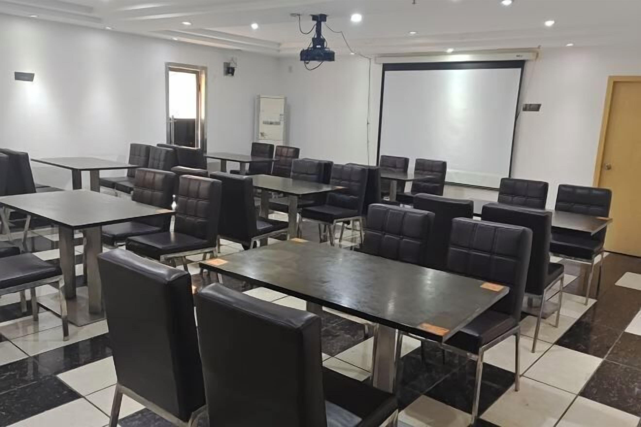 Un salón de conferencias con varias mesas y sillas, una pantalla para proyector y una pizarra.