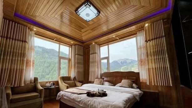 A bedroom with a large bed, two chairs, a nightstand, and a view of mountains through large windows. - Niegan Dawa Hotel (Diebu)