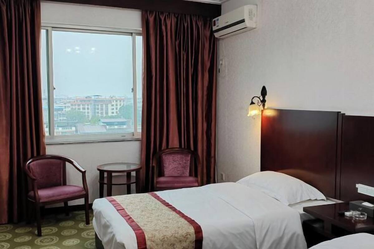 A hotel room with a bed, two chairs, a small table, an air conditioning unit, and a view of buildings through the window.