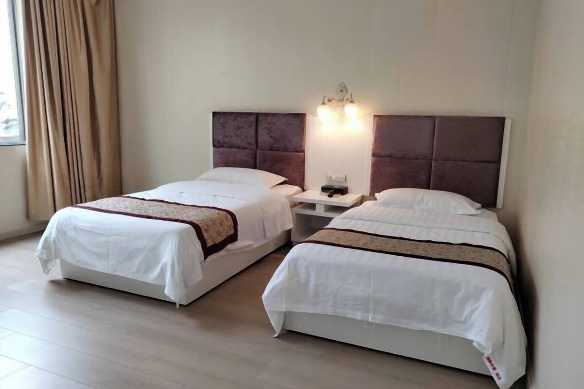 A hotel room with two beds, a nightstand, a lamp, and a window with curtains.