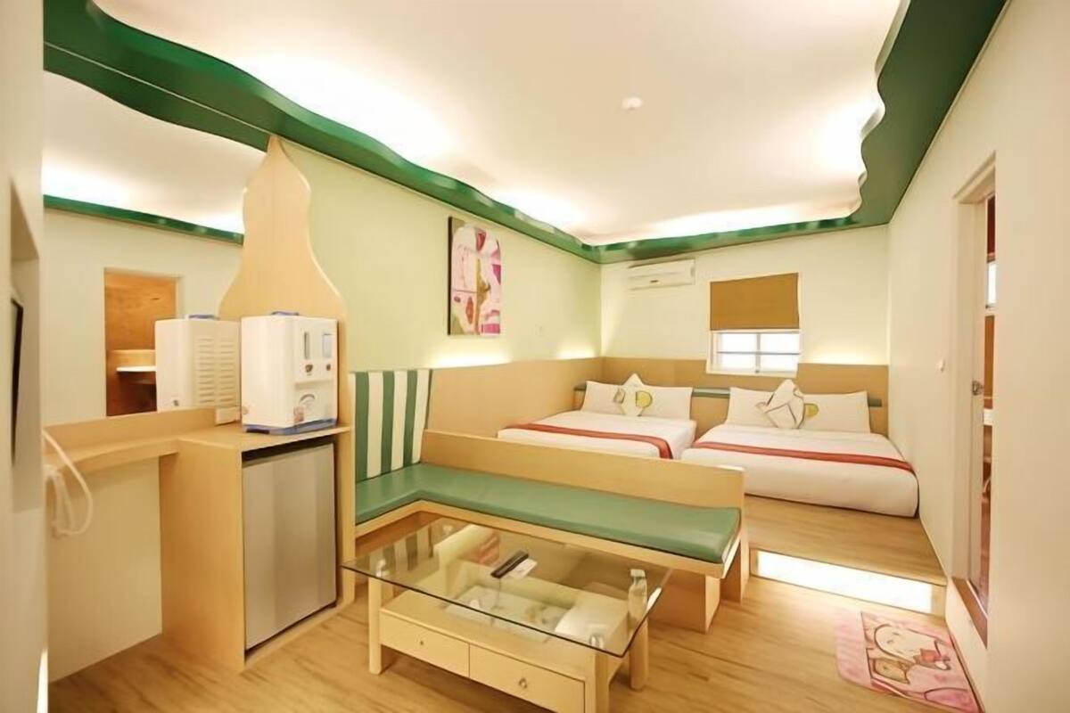 A hotel room with a kitchenette, a glass-top coffee table, two beds, and a green accent wall.