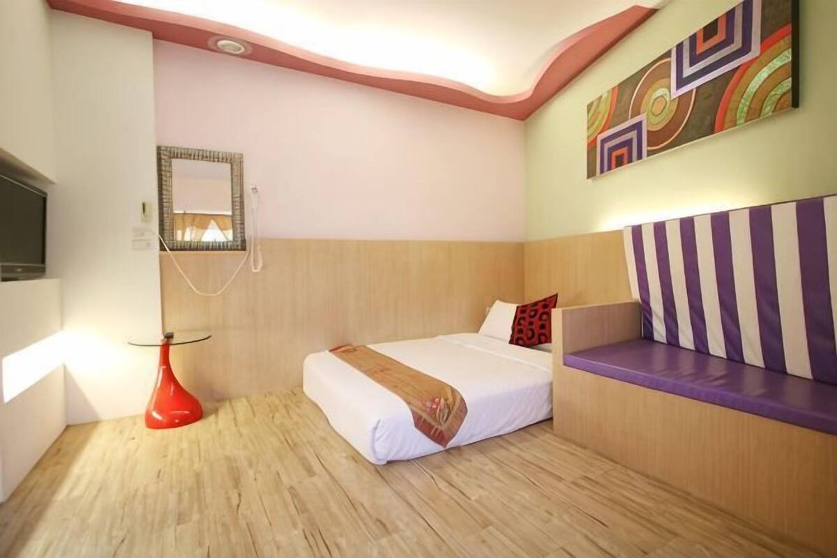 A hotel room with a bed, a bedside table, a mirror, a red lamp, and a colorful wall art piece.