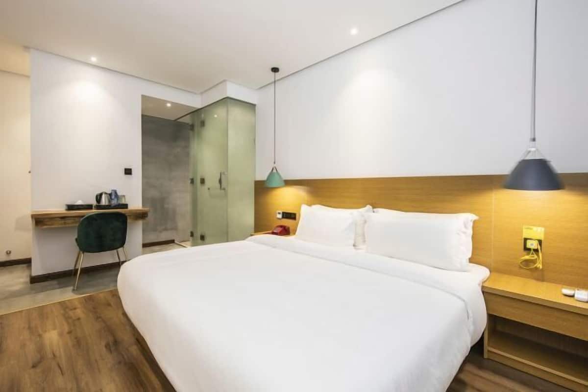 A modern hotel room with a large bed, a desk with a chair, a shower area, and a bedside table with a lamp.