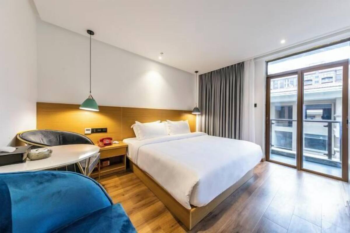 A hotel room with a bed, a desk, a chair, a small table, a lamp, and a large window with a view.