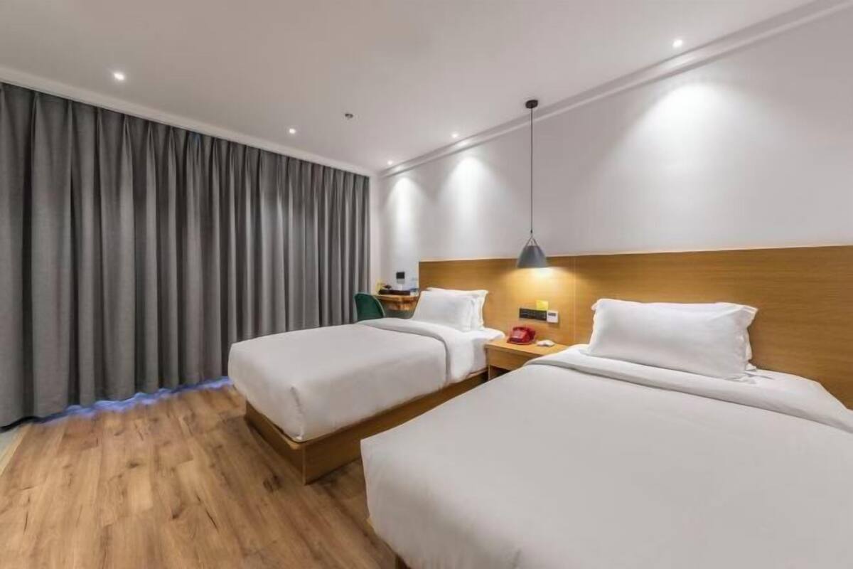 A hotel room with two beds, wooden flooring, a grey curtain, a bedside table with a lamp, and a wooden headboard.