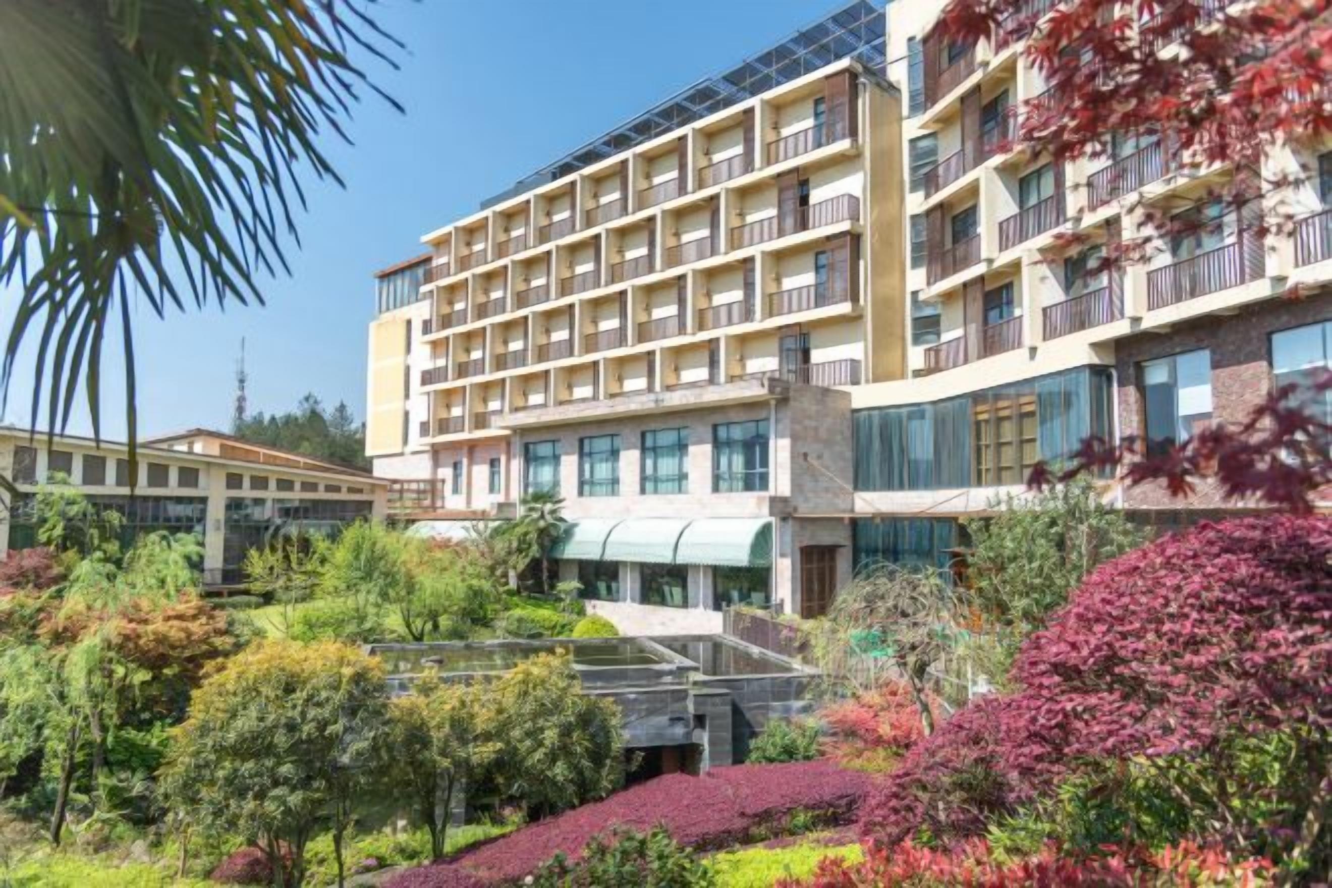 Un hotel de varios pisos con un diseño moderno, con balcones en cada planta, rodeado de jardines ajardinados y un cielo despejado.