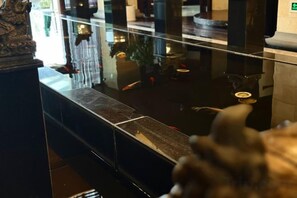 Una fuente reflectante con una estatua al frente, barandilla de vidrio y reflejo del interior de la habitación.