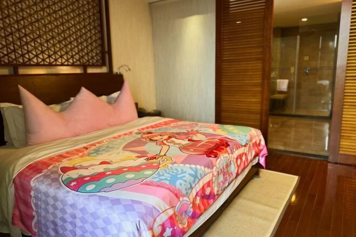 Cama com colcha colorida, cabeceira de madeira e vista para área de chuveiro com vidro.