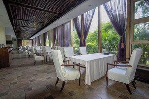 A dining area with tables and chairs, large windows with curtains, and a ceiling with a grid pattern.