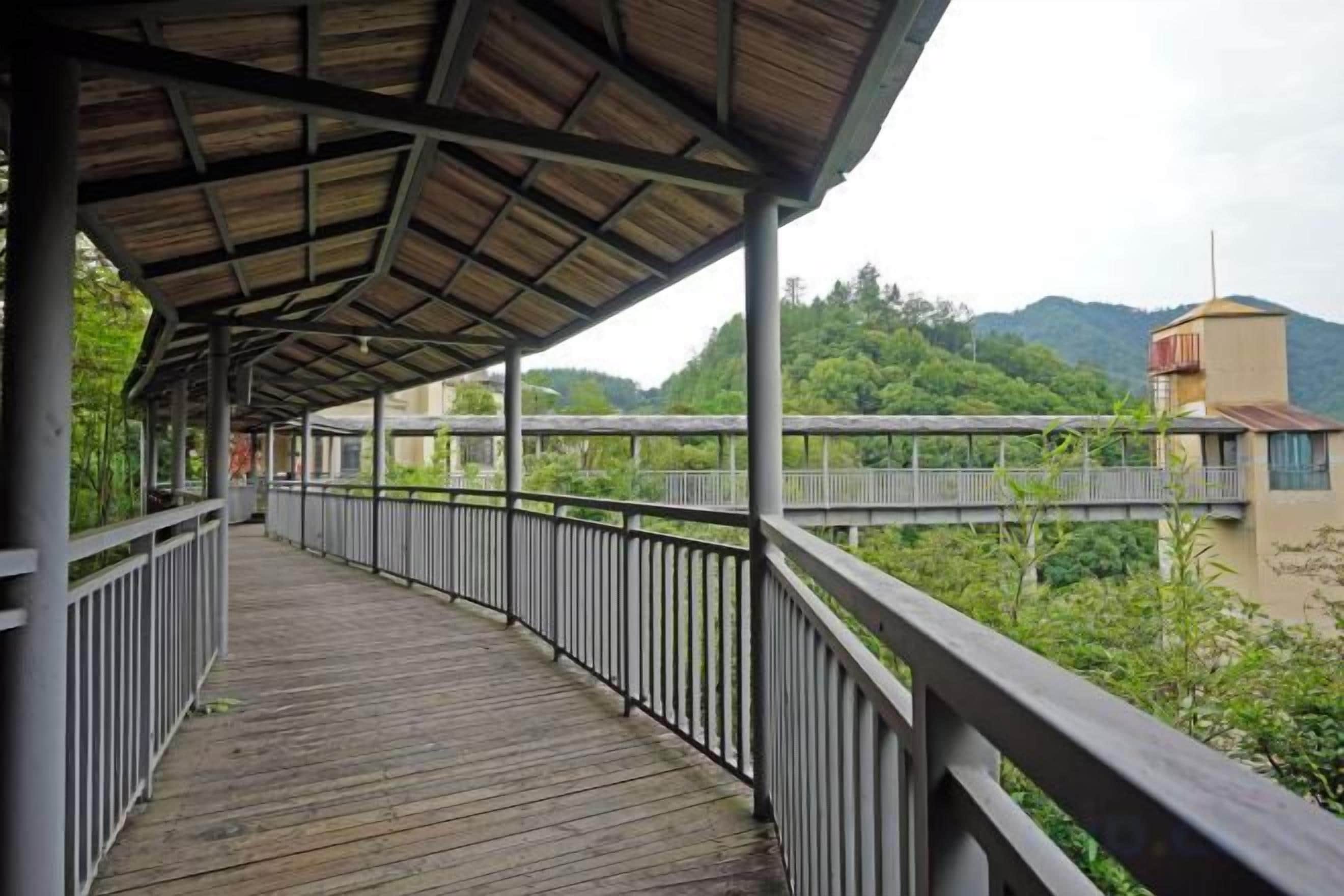 Une passerelle en bois couverte, dotée de rampes métalliques, qui mène à un pont enjambant une rivière, le tout étant entouré de verdure et de collines.