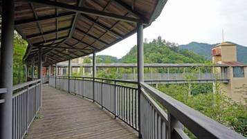 Une passerelle en bois couverte, dotée de rampes métalliques, qui mène à un pont enjambant une rivière, le tout étant entouré de verdure et de collines.