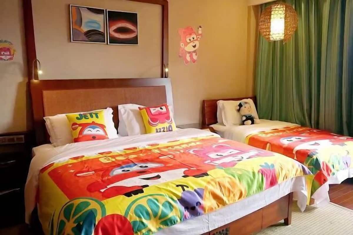 Quarto de hotel com duas camas, roupa de cama colorida, cabeceira de madeira, cortina verde e luminária pendente.