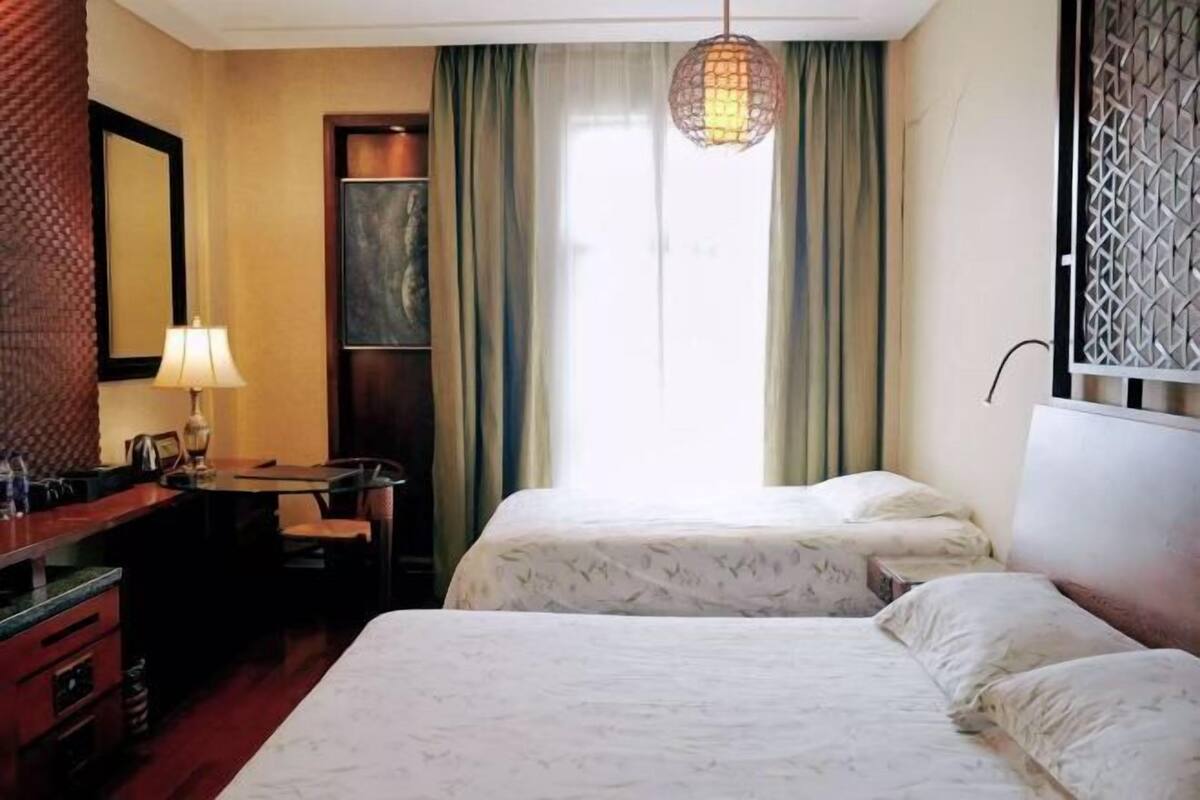 Quarto de hotel com duas camas, uma escrivaninha, uma cadeira, um abajur e uma janela com cortinas.