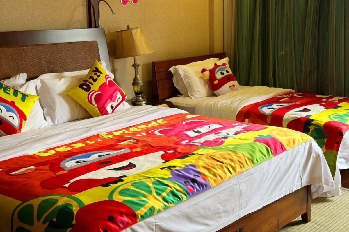 Quarto de hotel com duas camas, cada uma com roupa de cama colorida estampada com personagens de desenho animado. Há uma luminária na mesa de cabeceira entre as camas.