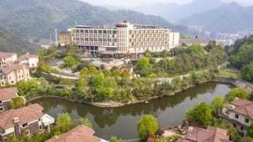 A hotel with a river in the foreground, surrounded by greenery and hills.