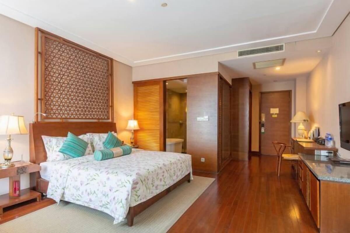 Quarto de hotel com cama, mesas de cabeceira, escrivaninha, televisão e porta de correr para um banheiro.