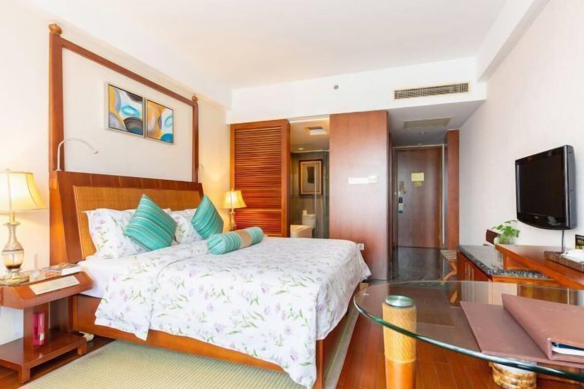 Quarto de hotel com cama, mesa de cabeceira, luminária, televisão e divisória de vidro.