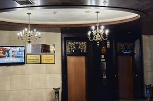 Um hall de hotel com lustres, uma televisão e dois elevadores.