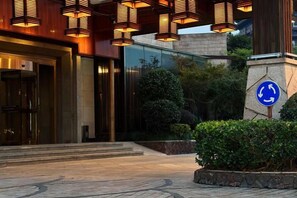 L’entrée d’un hôtel présente un design moderne, une façade en verre et un espace paysager agrémenté d’un panneau indiquant l’emplacement d’une poubelle de recyclage.