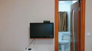 Una habitación con un televisor de pared, un estante blanco y un baño visible a través de una puerta abierta.