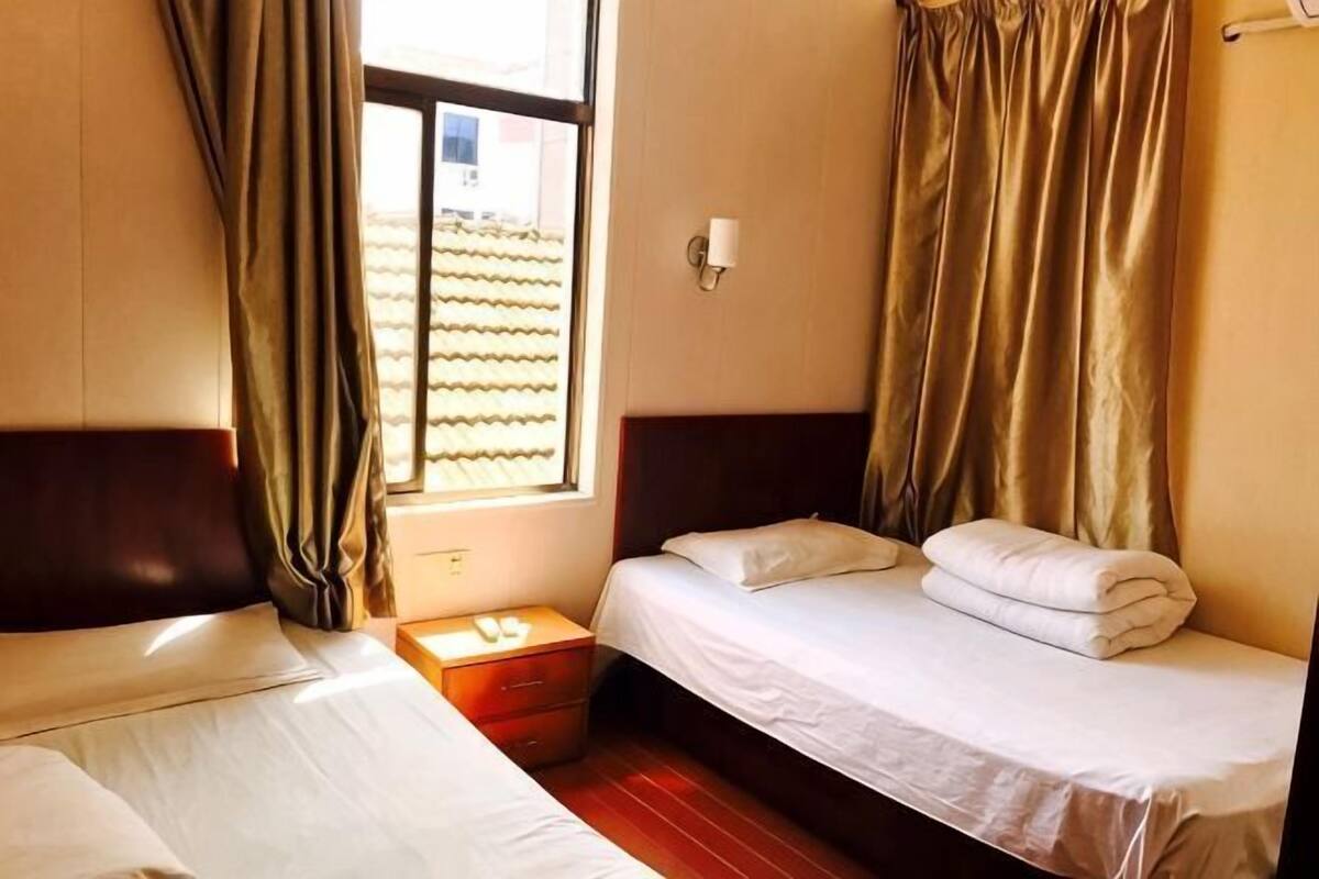 A room with two beds, a nightstand, a window with curtains, and a wall-mounted light.