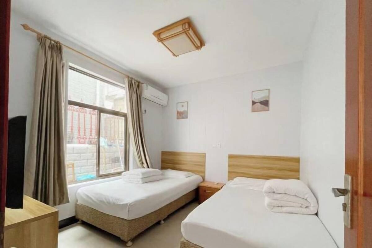 A hotel room with two beds, a window with curtains, a wooden headboard, a bedside table, and a wall-mounted air conditioner.