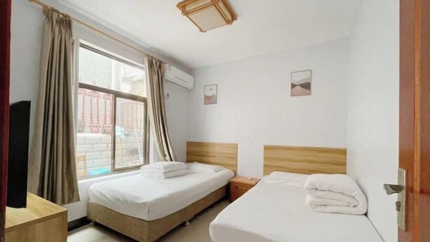 Habitación de hotel con dos camas, una ventana con cortinas, cabecero de madera, mesita de noche y aire acondicionado de pared.