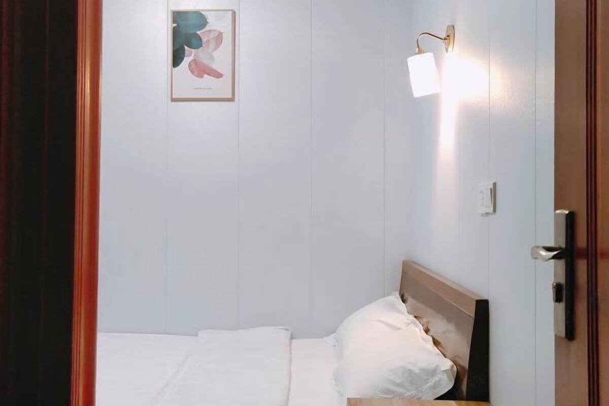 A single bed with white bedding, a wooden headboard, a wall-mounted light fixture, and a framed picture on the wall.