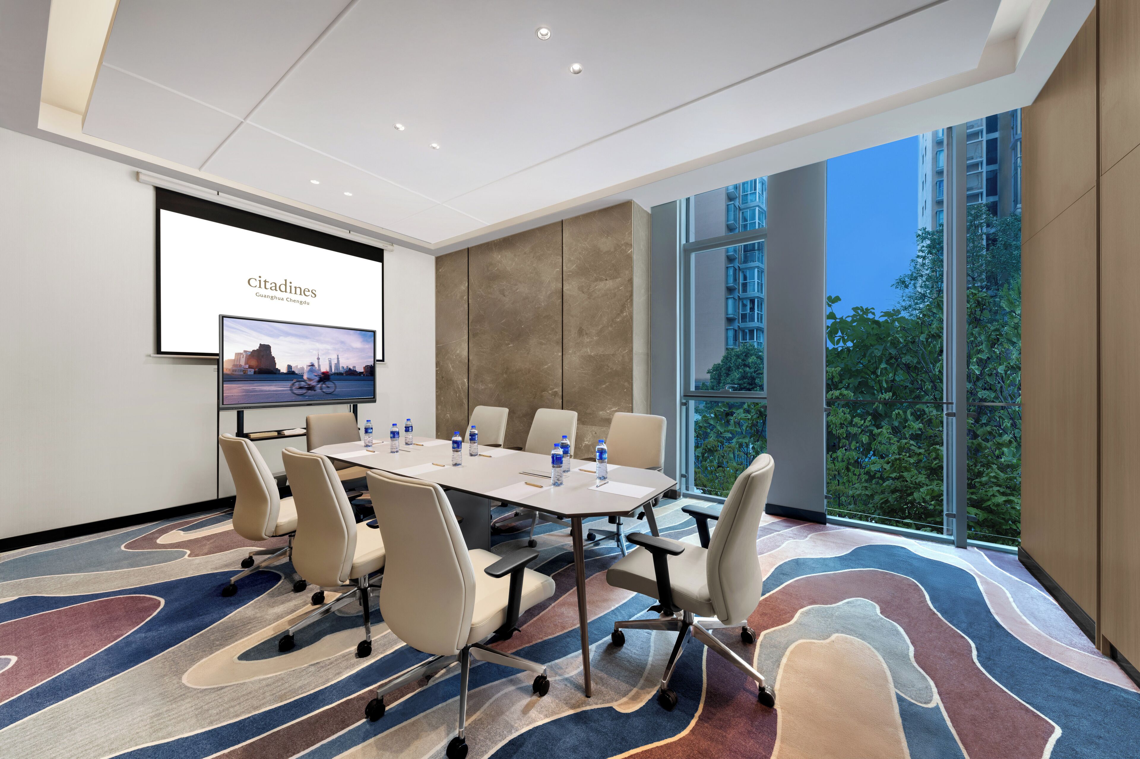 A modern conference room with a large table, comfortable chairs, a projector screen, and a large window with a city view.