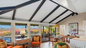 Habitación amplia con techo alto, ventanas grandes y vista a un jardín. Se observan muebles de mimbre, una mesa de comedor y un sofá.