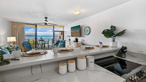 Een moderne woonkamer met een groot raam met uitzicht op de oceaan, een keukenhoek met fornuis, en een eethoek met blauwe stoelen.