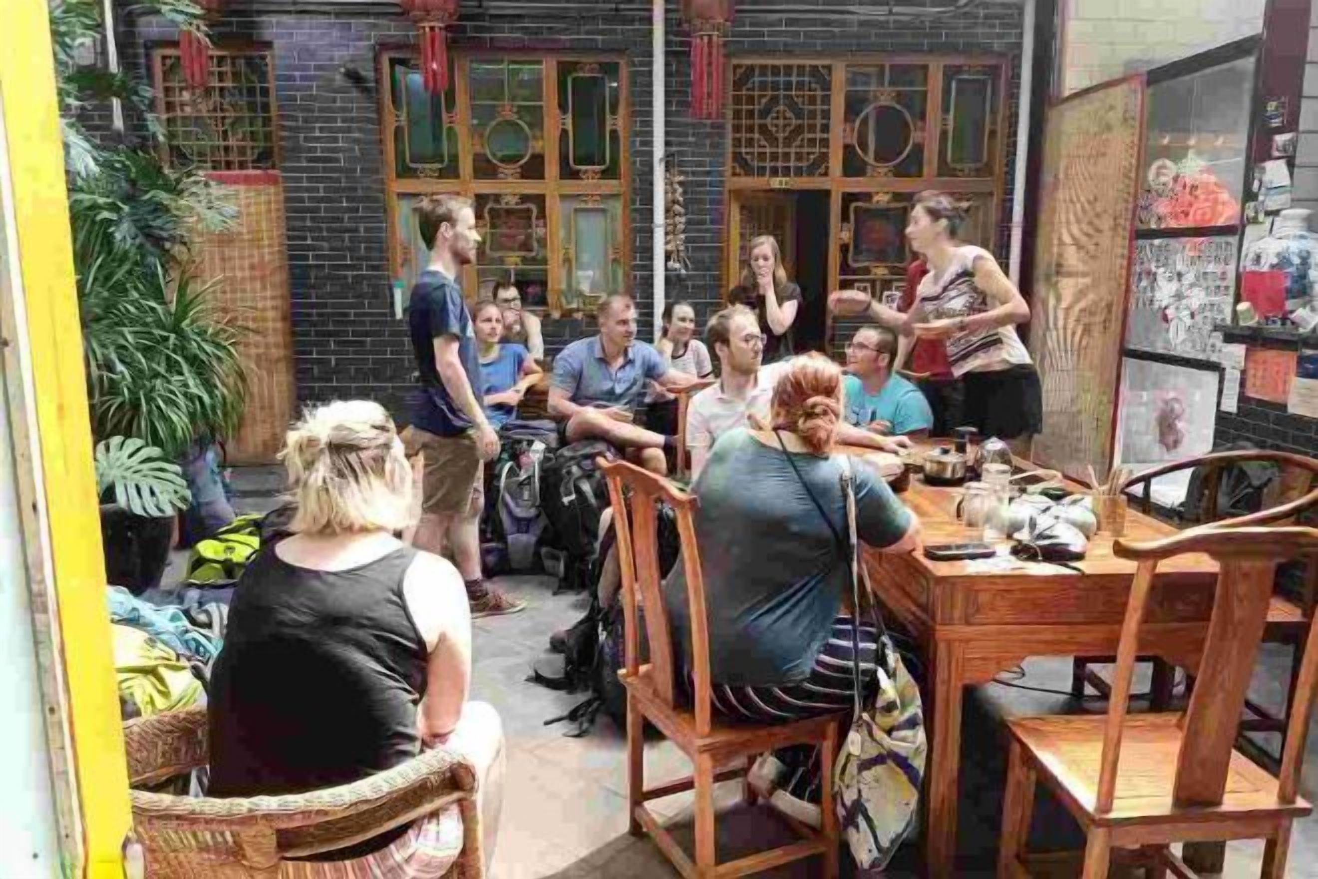 A group of people seated around a wooden table in an outdoor setting with brick walls and potted plants.