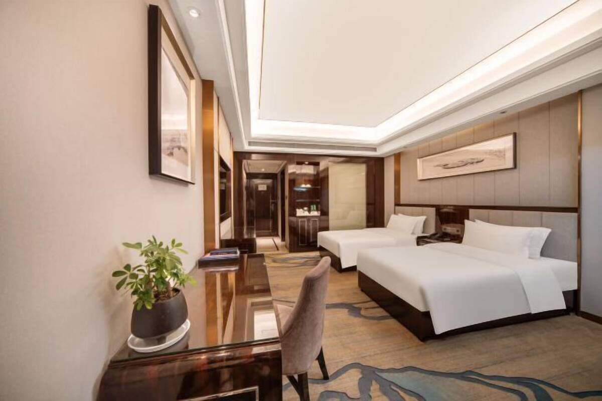 A hotel room with a bed, a desk, a chair, a potted plant, and a framed picture on the wall.