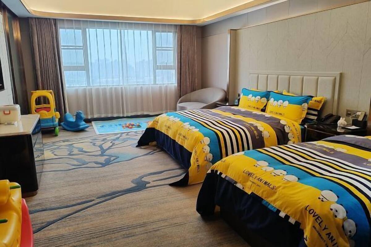 A hotel room with two beds, a desk, a chair, a window with curtains, and a view of the city.