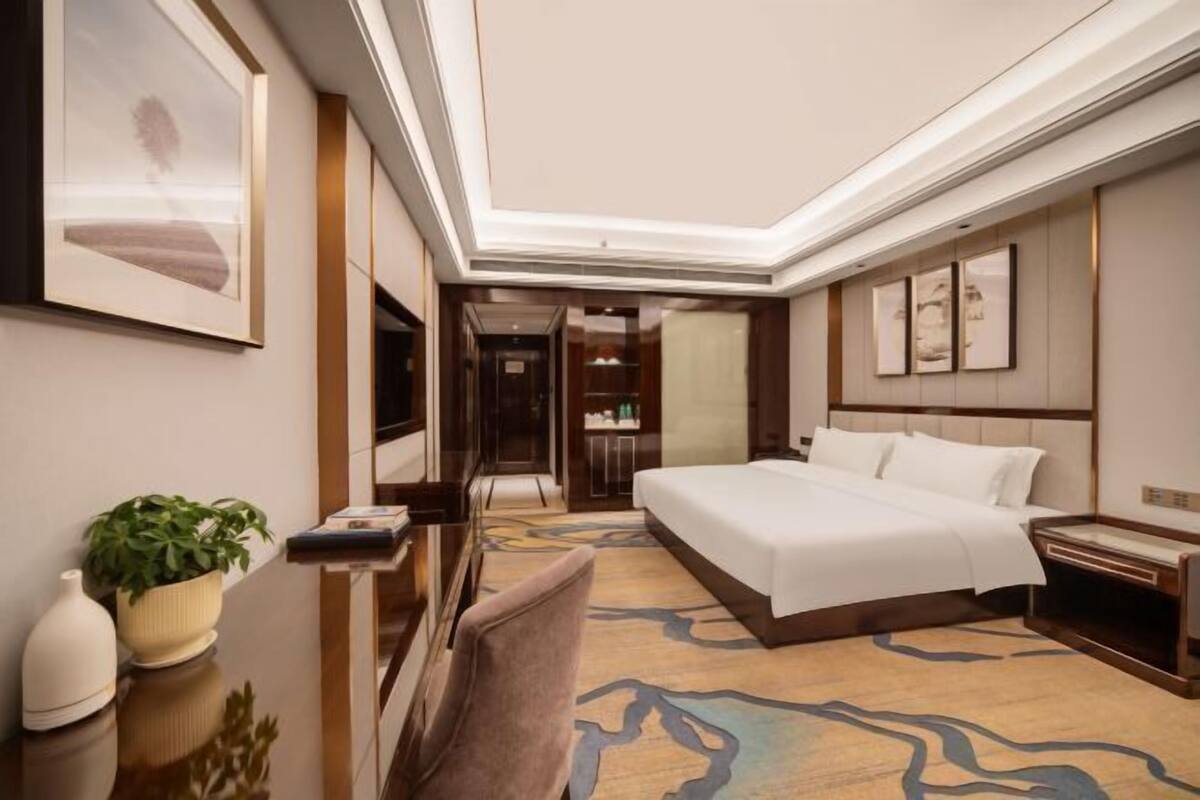 A hotel room with a large bed, a bedside table, a chair, a plant, and framed artwork on the walls.