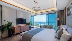 A modern hotel room with a large bed, a TV, a wooden cabinet, and a balcony with a view of the ocean.