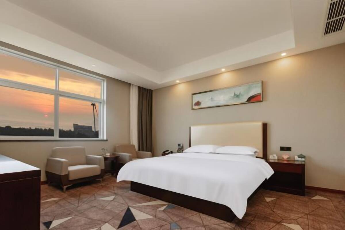 Habitación de hotel con cama, escritorio, silla, mesita de noche, un cuadro y un ventanal con vistas a la puesta de sol.