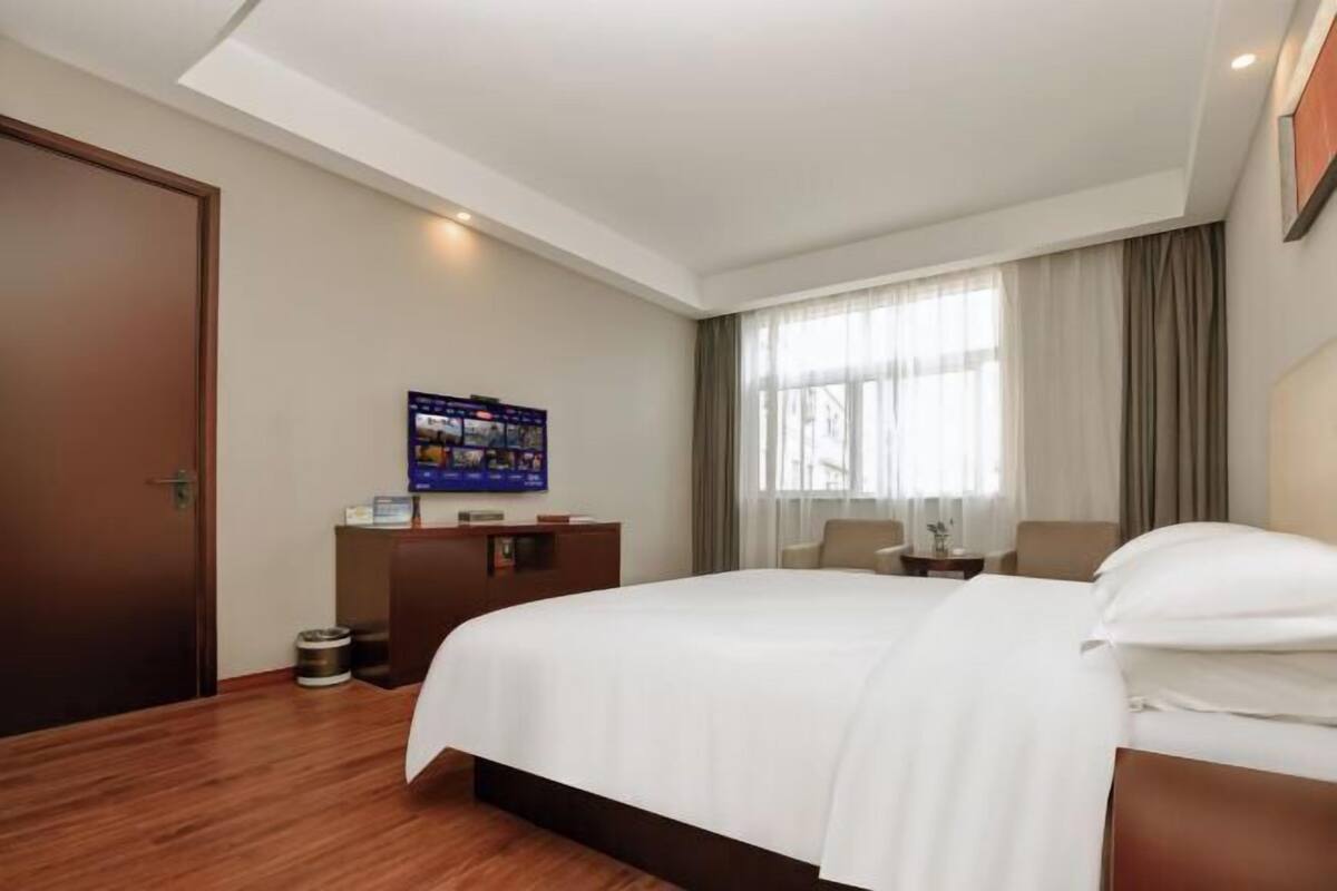Habitación de hotel con una cama grande, piso de madera, un escritorio con computadora, televisor y ventana con cortinas.