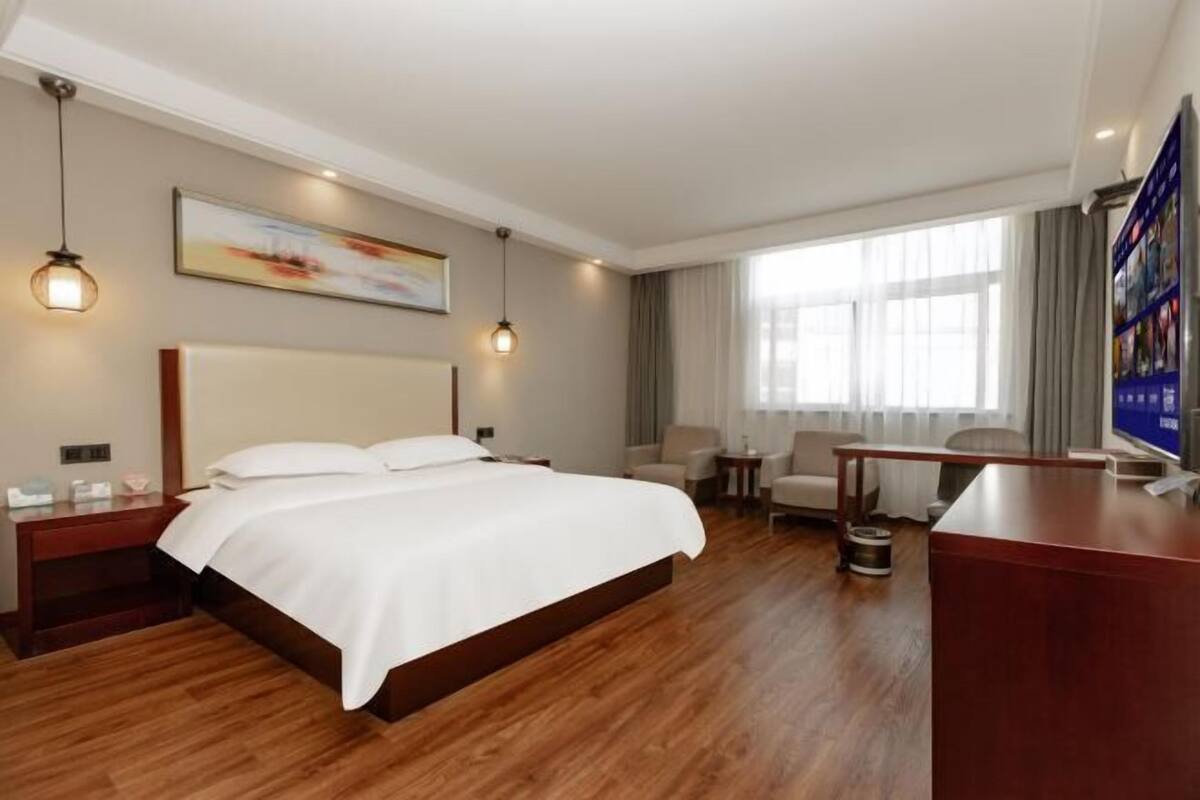 Habitación de hotel con una cama grande, mesitas de noche, un escritorio, una silla, un televisor y una ventana con cortinas.